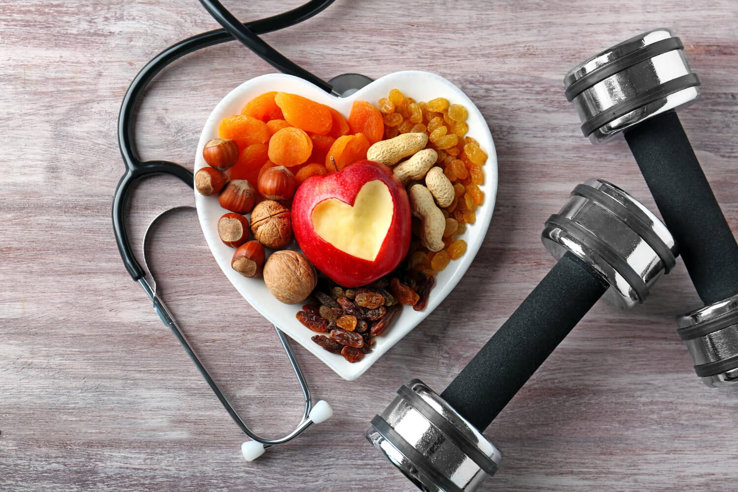 Heart-shaped white bowl filled with healthy foods including dried apricots, golden raisins, peanuts, hazelnuts, walnuts, and a red apple with a heart-shaped cutout. A stethoscope lies beside the bowl, and two metallic dumbbells with black handles are placed to the right on a wooden surface, symbolizing heart health and wellness through nutrition and exercise.