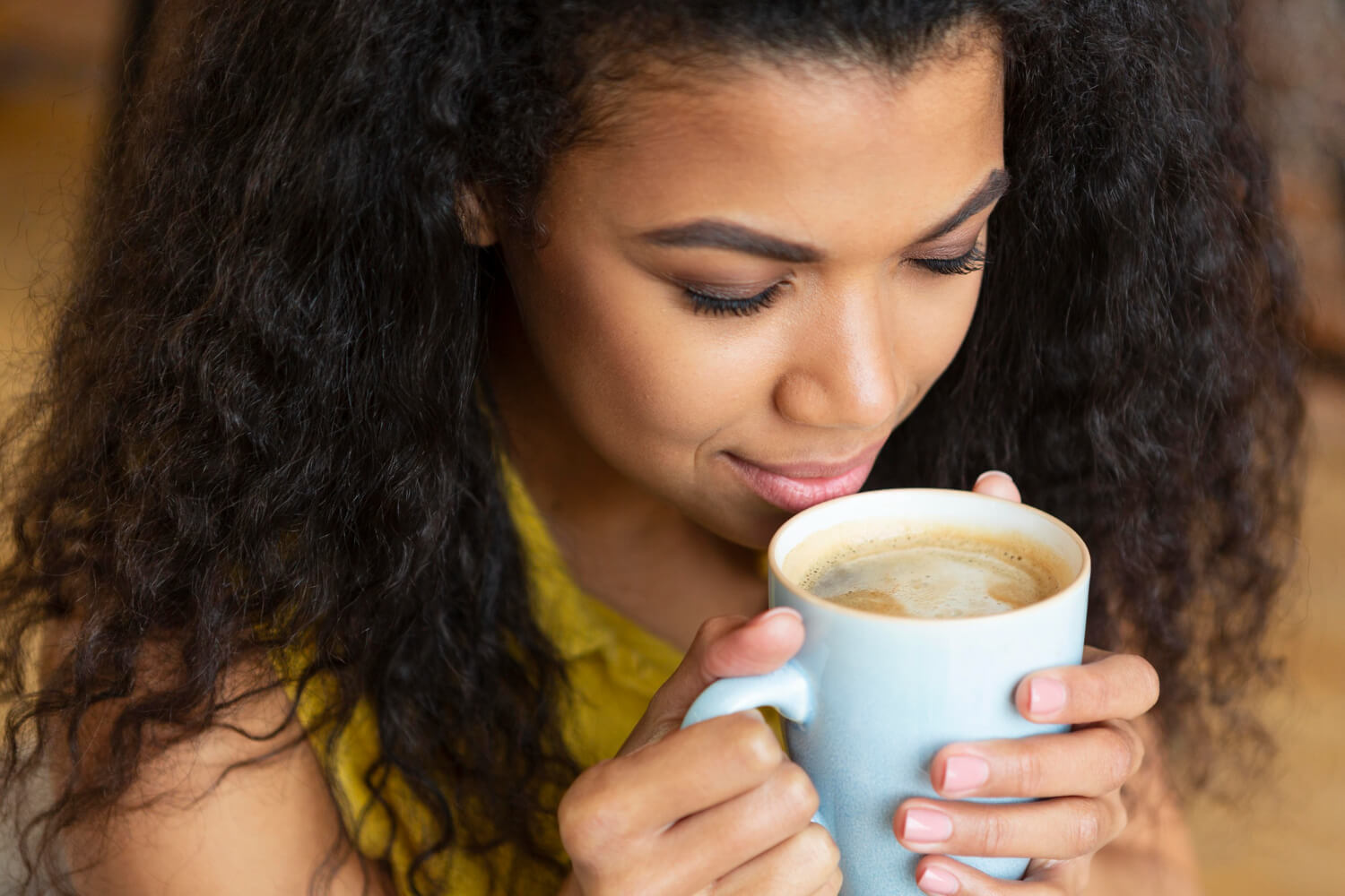 A close-up portrait of a young woman with curly black hair holding a light blue mug with both hands. She has her eyes gently closed and a subtle smile as she leans in to smell or sip her hot coffee.