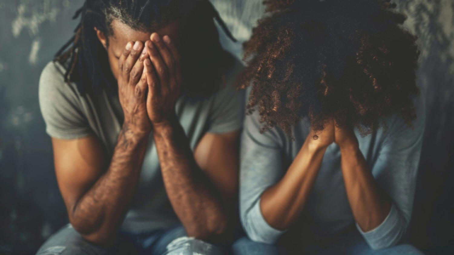 A Black man and woman sitting side by side, both resting their faces in their hands in distress.