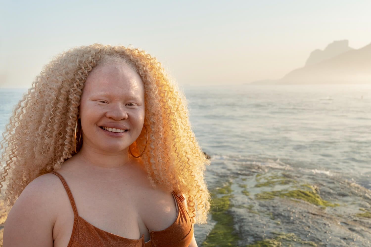 A portrait of a smiling woman with albinism wearing a terracotta top and hoop earrings, standing near a calm ocean shore. Her long, voluminous golden-blonde curls are lit by soft sunlight, with hazy mountains and water in the background.